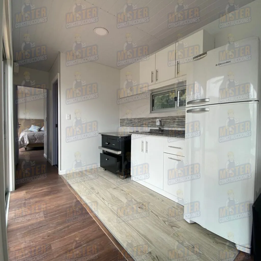 Cocina moderna con muebles blancos, refrigerador y horno, vista hacia un dormitorio con cama en fondo